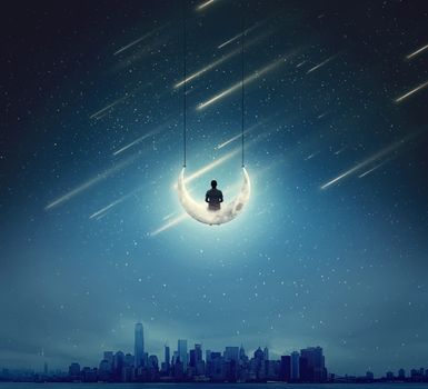 Surreal background with a boy sitting on a crescent moon, as a swing, over a big city in a starry night
