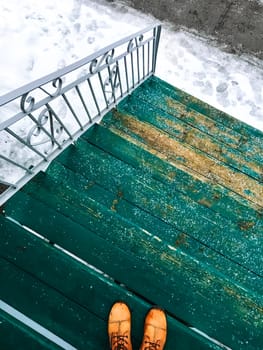 Person in orange boots standing on a green wooden staircase leading onto a winter street.