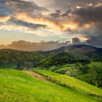 summer landscape. fence near the meadow path to village on the hillside. forest with lumber on the mountain at sunrise