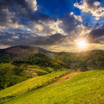 summer landscape. fence near the meadow path to village on the hillside. forest with lumber on the mountain at sunset