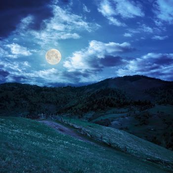 summer landscape. fence near the meadow path to village on the hillside. forest with lumber on the mountain at night in full moon light