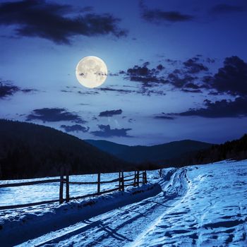 winter mountain landscape. winding road that leads into the pine forest covered with snow. wooden fence stands near the road at night in full moon light