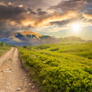 gravel road going off into the distance and passes through the green field in mountains at sunset