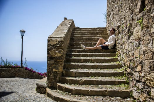 Side view of stylish man in sunglasses sittingon stone stairs and relaxing. Ocean on background.