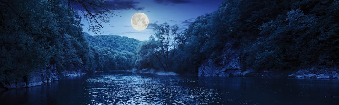 mountain river with stones on the shore in the forest near the mountain slope at night in full moon light
