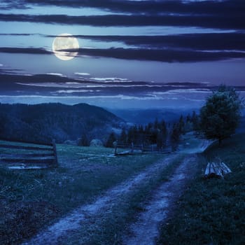 fence and lonely tree near the path through meadow  on the hillside. forest in fog on the far hill and mountain at night in full moon light