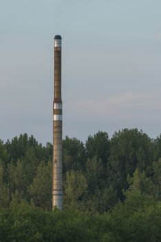 Factory chimney of a factory located in a forested area visible from a nearby higher point in the evening light
