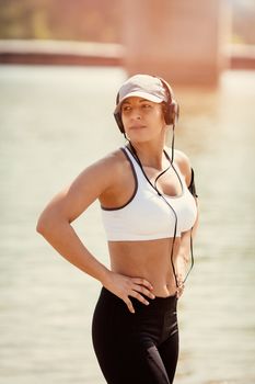 Young muscular woman with headphones resting after jogging by the river and looking away.