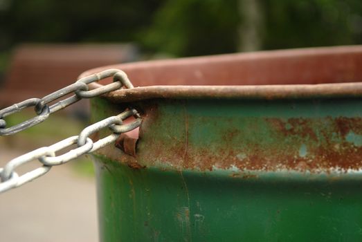 Image of a chained rusted garbage can in a park. Selective focus on where the chain is connected to the garbage can. Lots of space for content.