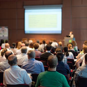 Business and entrepreneurship symposium. Speaker giving a talk at business meeting. Audience in conference hall. Rear view of unrecognized participant in audience.