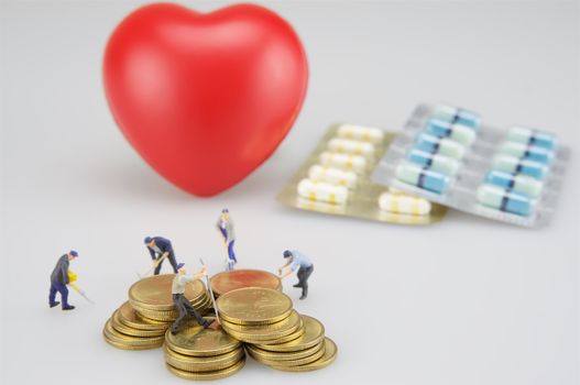 Pile of coins with miniature people, packing of medicine and red heart.