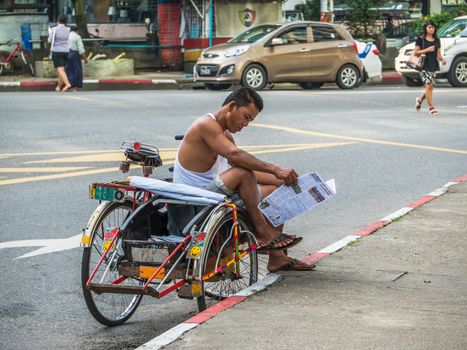 Yangon - September 9, 2016: Tricycle taxis are still common and a practical way of transportation in Myanmar’s capital Yangon. Here’s a taxi driver who is reading a newspaper while waiting for customers.