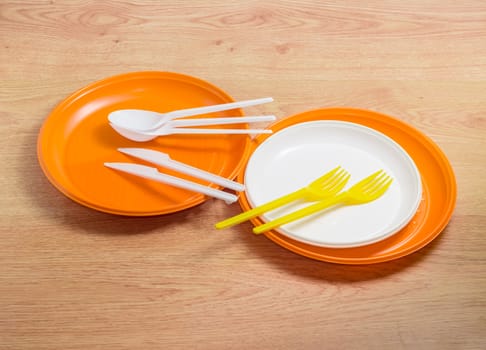 Orange and white disposable plastic plates different sizes, plastic disposable yellow forks and white spoons and knives on a wooden surface
