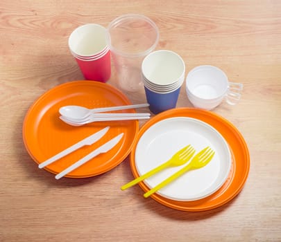 Orange and white disposable plastic plates different sizes, plastic disposable forks, spoons and knives, disposable plastic and paper cups on a wooden surface
