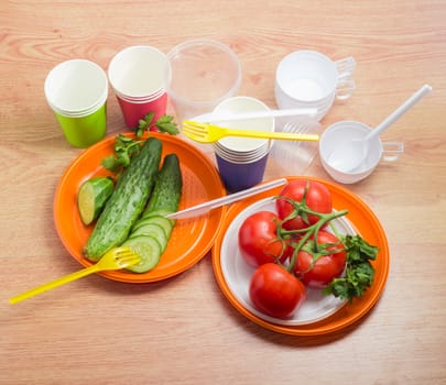 Different disposable plastic plates, partly sliced cucumbers with fork and knife and cluster of the tomatoes on them, paper and plastic disposable cups with spoons beside on a wooden surface
