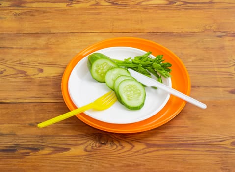 Pile of several white and orange disposable plastic plates different sizes, sliced cucumber and parsley twig with disposable plastic fork and knife on it on a surface of the old wooden planks
