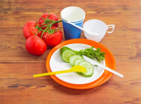 Disposable plastic plates, fork, knife and sliced cucumber on it, claster of the tomatoes, paper and plastic disposable cups with spoons beside on a surface of the old wooden planks
