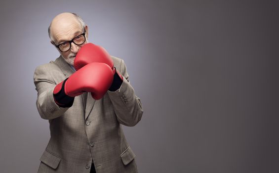 Senior man making hit wearing boxing gloves on gray background