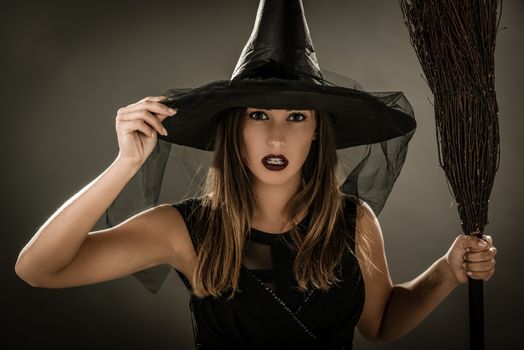 Portrait of a young woman dressed like a witch. She is in dark clothing and holding a broom. Looking at camera.