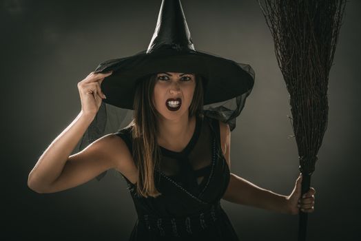 Portrait of a young woman dressed like a witch. She is in dark clothing and holding a broom. Looking at camera.