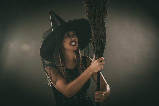 Young woman dressed like a witch. She is in dark clothing and holding a broom. 