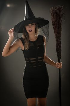 Young woman dressed like a witch. She is in dark clothing and holding a broom. Looking at camera.