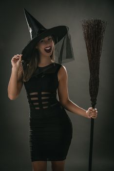 Young woman dressed like a witch. She is in dark clothing and holding a broom. Looking at camera.