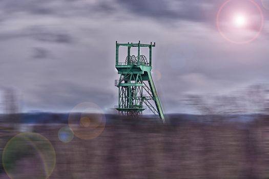 Image with lens flare effect and blur: Age headframe industrial monument in the Ruhr area before dawn sky with clouds.