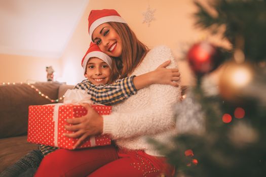 Beautiful young smiling mother give a present to her cute happy son for a Christmas or New Year at the home. Looking at camera.