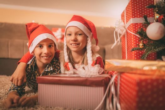 Cute smiling sister and brother with many presents at the home in a Christmas time. They are embraced and looking at camera.