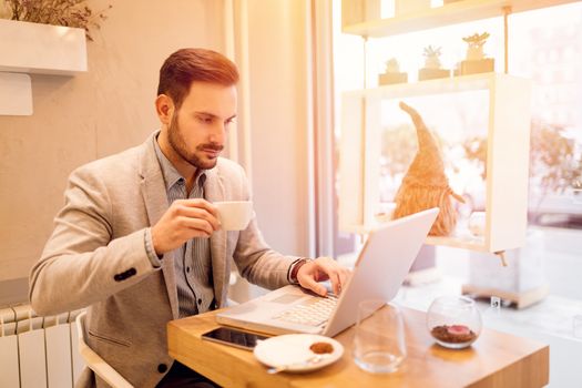 Young smiling businessman on a break in a cafe. He is drinking coffe and working at laptop. 