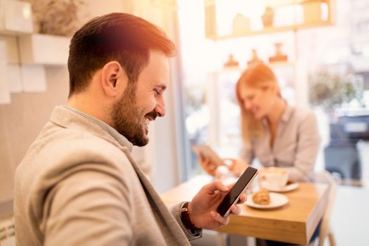 Young smiling businesspeople on a break in a cafe. Man using smart phone and reading message from internet. Woman working at tablet. Selective focus. Focus on foreground, on businessman.