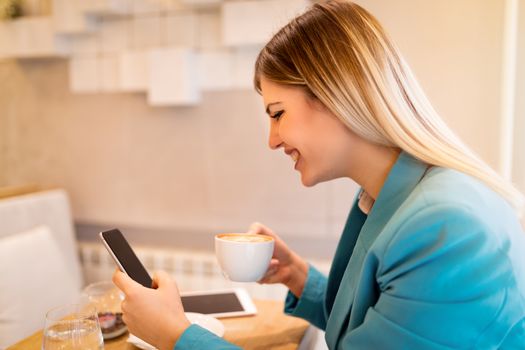 Young smiling businesswoman on a break in a cafe. He is drinking coffee and reading message on smart phone. 