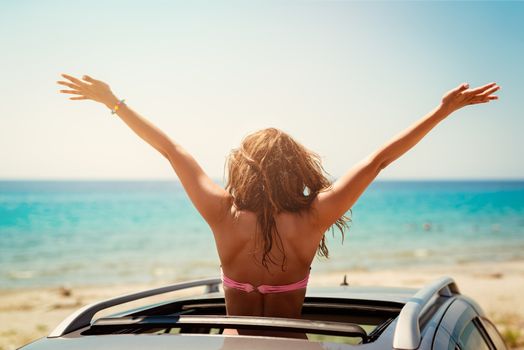 Rear view of a beautiful young woman posing with raised hands in a cabriolet on the beach.