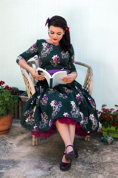 Vintage girl with beautiful floral dress reading a book on a wicker chair.