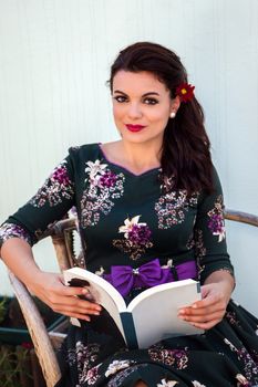 Vintage girl with beautiful floral dress reading a book on a wicker chair.