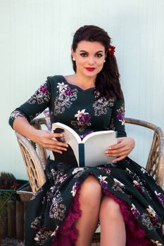 Vintage girl with beautiful floral dress reading a book on a wicker chair.