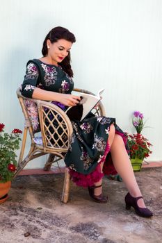 Vintage girl with beautiful floral dress reading a book on a wicker chair.