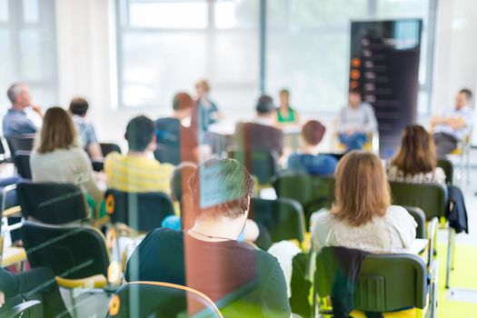 Round table discussion at business and entrepreneurship symposium. Audience in conference hall. Lens focus on unrecognized participant in rear of audience. Shot trough glass.