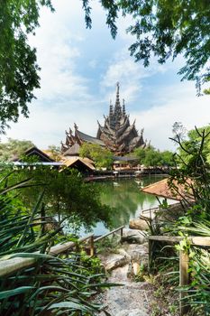 Path leading towards the river through tropical trees. In the background large wooden temple and blue sky. Pattaya, Thailand