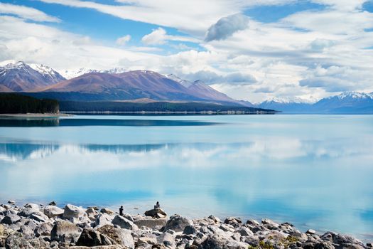 Girl sits on large rock while man takes photo. Lake Pukaki in New Zealand in background surrounded by mountains.