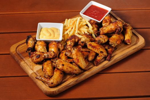 big wooden board with grilled chicken wings with french fries and two sauces, preferably served as companion for beer or other alcohol drinks