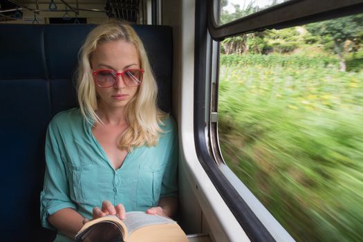 Blonde caucasian woman reading book by train window , traveling by train in third world country. Blured green fields can be seen trough window.