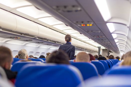 Interior of airplane with passengers on seats and stewardess walking the aisle.