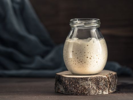 Active wheat sourdough starter in glass jar on brown wooden background. Starter for sourdough bread. Toned image. Copy space.