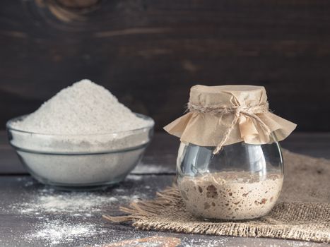 Active rye sourdough starter in glass jar and rye flour on brown wooden background. Starter for sourdough bread. Toned image. Copy space.