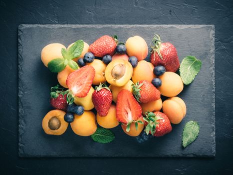 Top view of fruits and berries on dark stone plate. Heap of fresh strawberries, blueberries, apricot and mint leaves on black slate board. Mix of berries, healthy food, superfood, diet, detox concept.