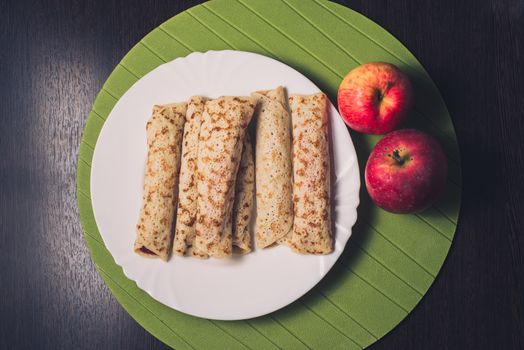 Roasted rolled pancakes on a white round plate and two apples near. Top view . Flat lay.