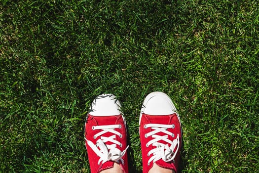 Legs in old red sneakers on green grass. View from above. The concept of youth, spring and freedom.