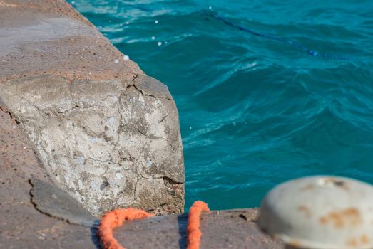 Photo of beautiful clear turquoise sea ocean water surface with ripples and bright splash on stone seascape background. Orange rope at pier.